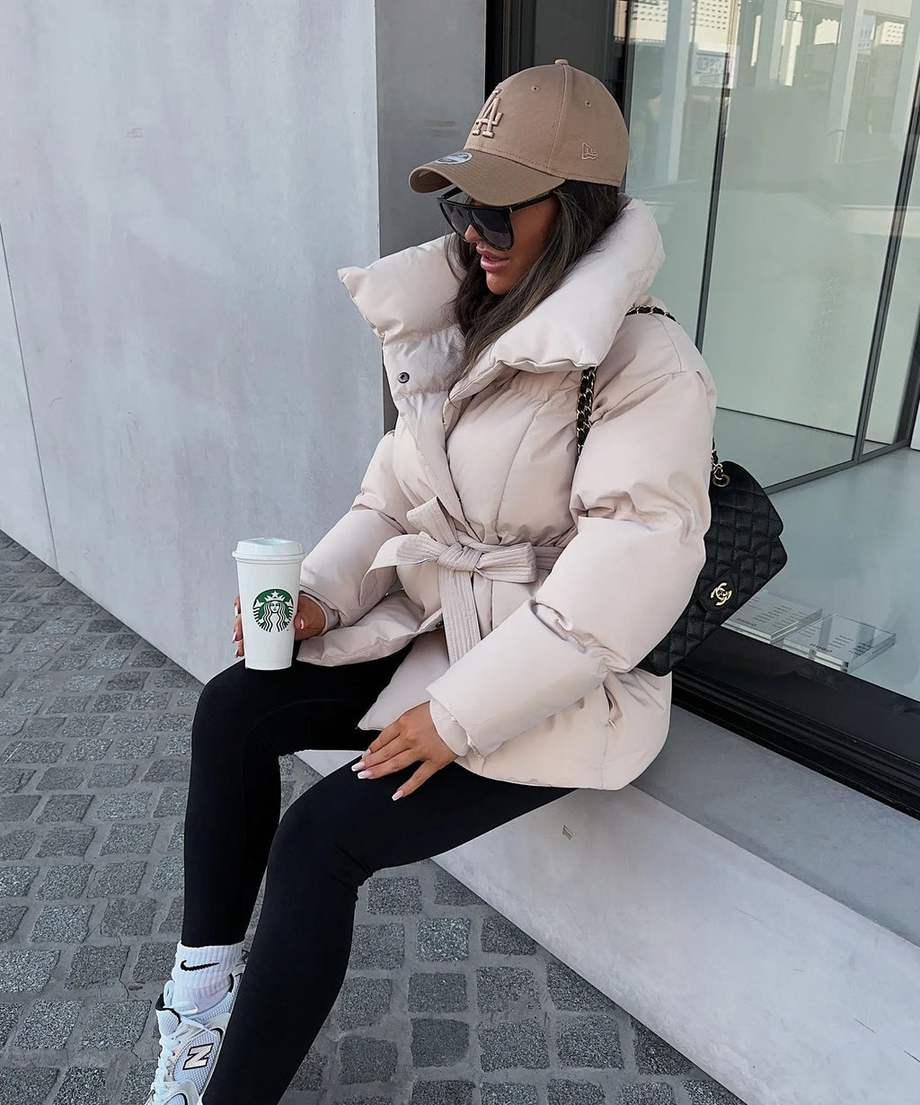 Woman seated wearing a cream puffer jacket tied at the waist, paired with black leggings and trainers