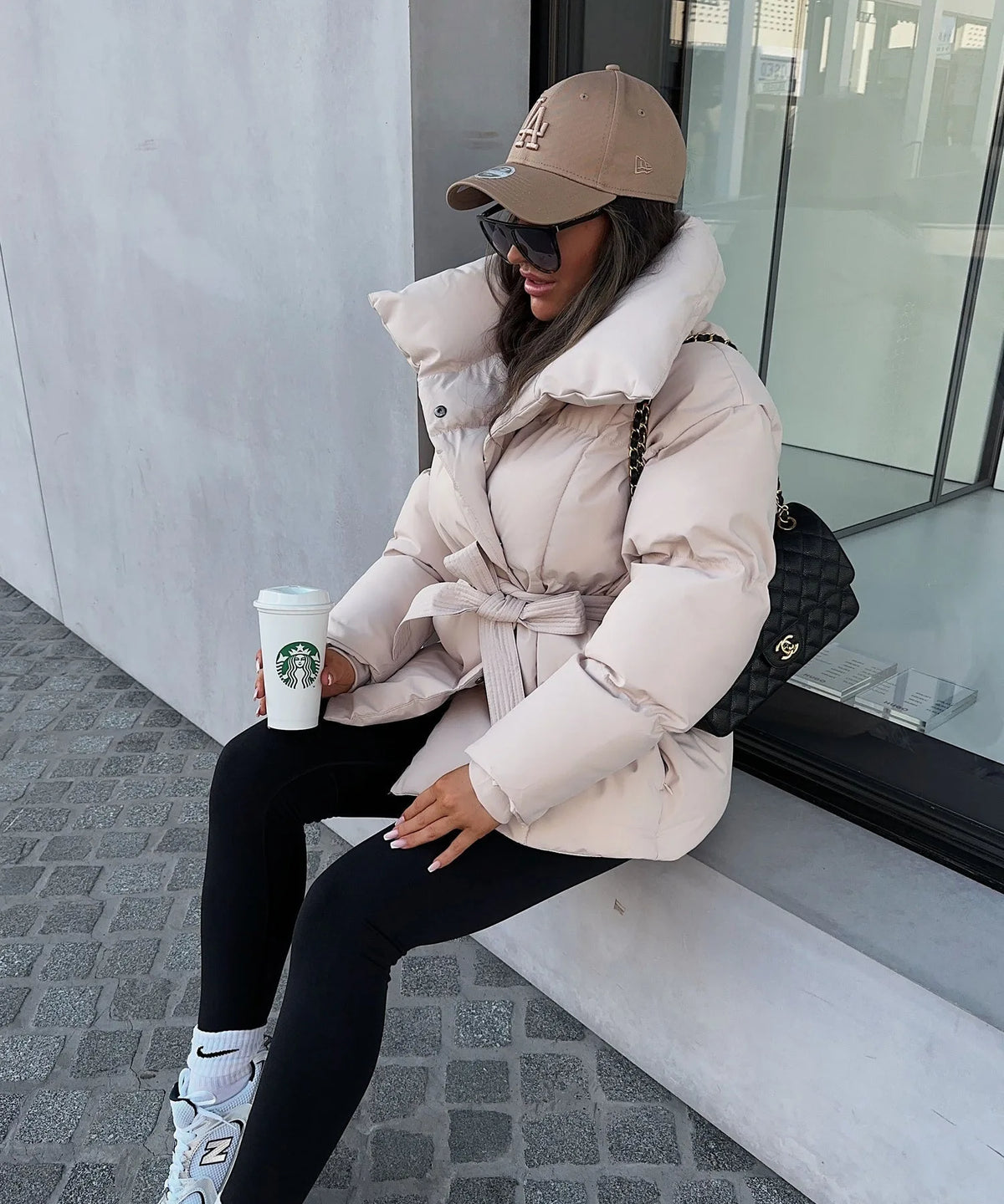 Woman seated wearing a cream puffer jacket tied at the waist, paired with black leggings and trainers