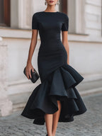 Front view of a woman in a black fitted midi dress with a high neckline and layered ruffle hem, holding a beaded clutch