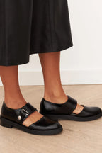 Side view of black leather Mary Jane flats with buckle detail, worn with cropped black trousers