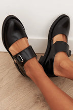 Woman resting bare feet showing both black buckle loafers with chunky soles against a white wall