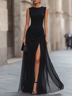 Black sleeveless maxi evening dress with ruched bodice and sheer high-slit skirt, worn on a city street.