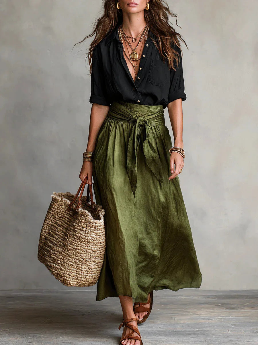 Woman in a black button-up shirt and olive green tie-waist midi skirt holding a woven straw tote bag
