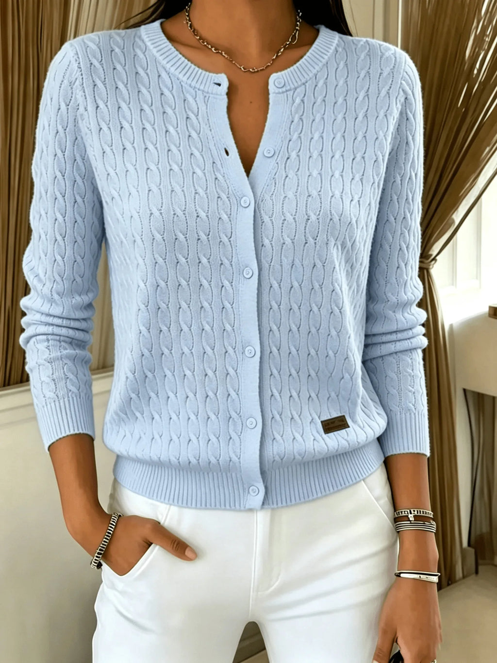 Light blue cable knit cardigan with buttons undone at the neckline, worn with white trousers and a necklace