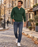 Man wears a forest green cable-knit Henley jumper over a white tee with dark jeans and white trainers on a cobbled street.