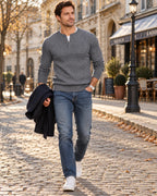 Grey cable-knit Henley jumper worn with a white tee, navy jeans and white trainers while carrying a dark overcoat.