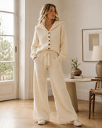 Woman wearing a cream ribbed knit hooded cardigan with wooden buttons and matching wide-leg knit trousers, standing by a window.