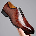 Angled close-up of a hand holding a dark brown croc-embossed slip-on loafer showing the textured leather and tan lining
