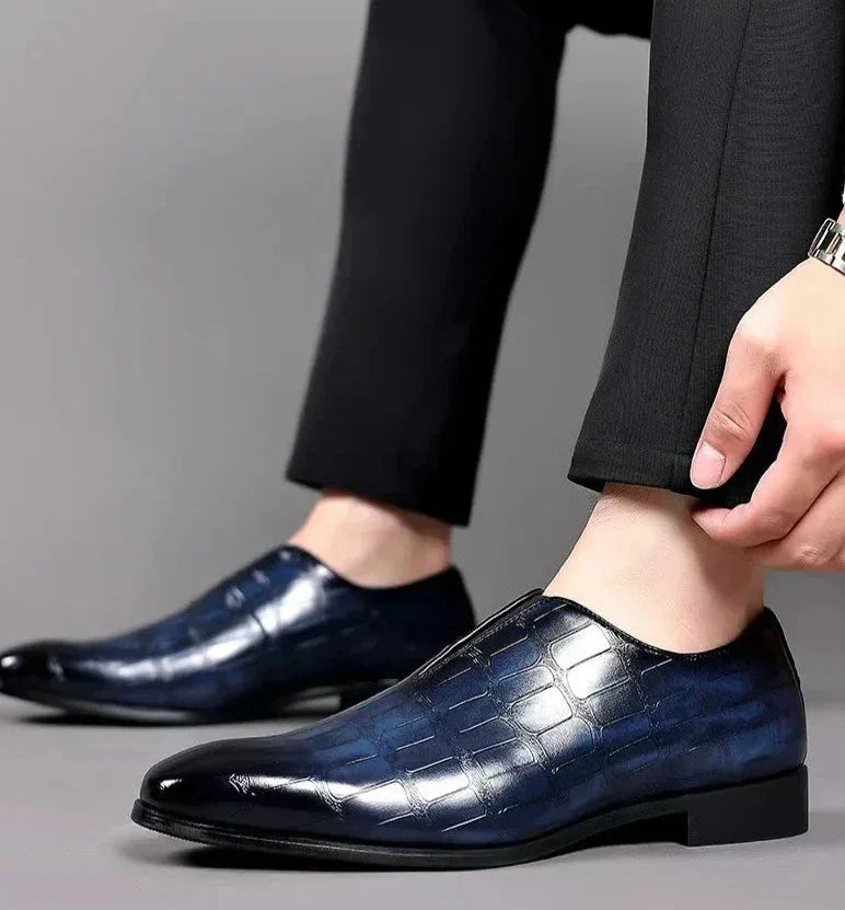 Close-up of a model's ankle wearing navy blue croc-embossed slip-on loafers with dark trousers