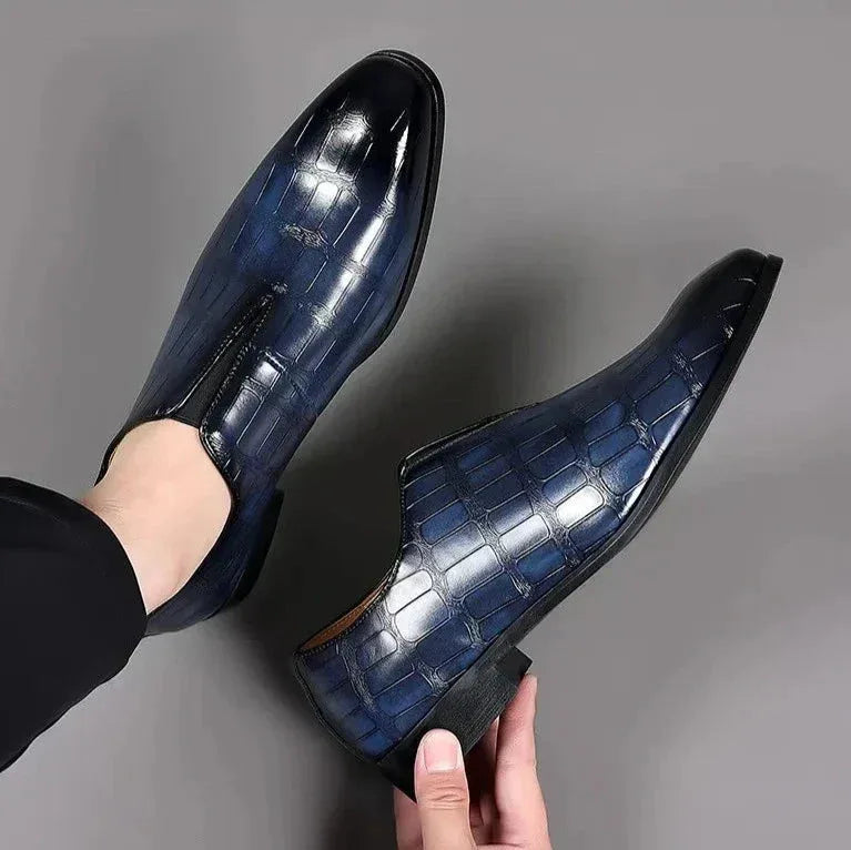Overhead view of a pair of navy blue croc-textured loafers held mid-air, showing the pointed toe shape