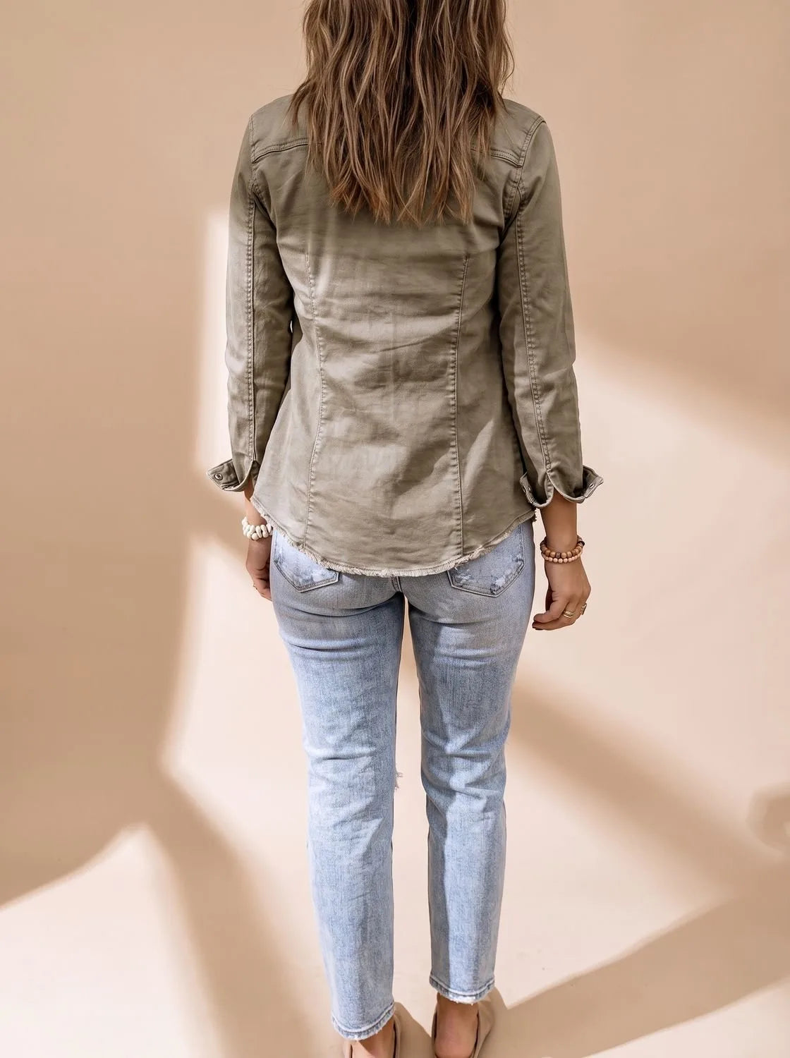 Back view of the olive green denim jacket showing its slightly fitted shape and raw hem paired with light wash jeans