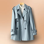 Pastel blue double-breasted trench coat with belted cuffs, shown on a wooden hanger against a peach backdrop.