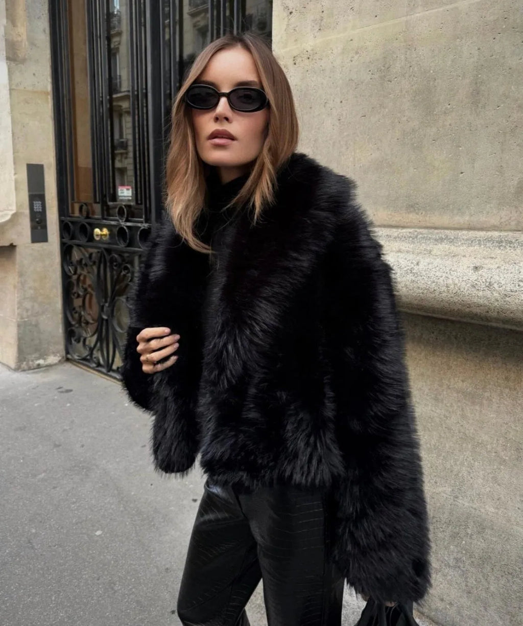 Woman wearing a cropped black faux fur coat with wide collar over black leather trousers on a Paris street