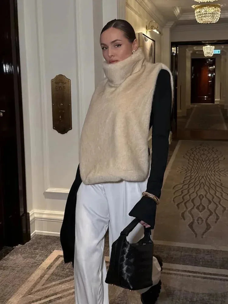Model wearing a beige faux fur gilet with high polo neck over a white long-sleeve top and white trousers
