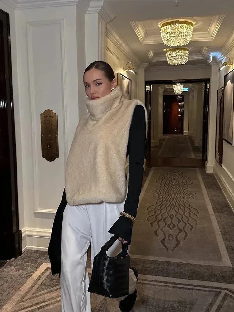 Front view of a beige faux fur cropped gilet with high polo neck styled over a black top and white trousers