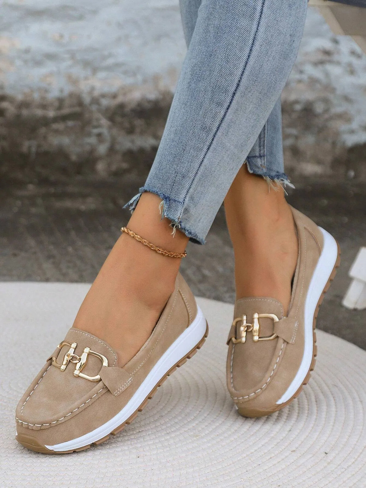 Beige suede loafer with gold horsebit buckle and ridged white and tan sole on straw mat
