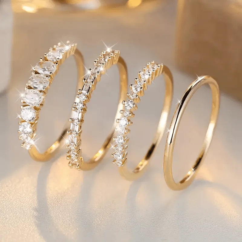 Four stacked gold-tone rings with clear zirconia stones shown side by side against a warm blurred backdrop