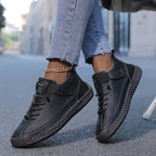 Side view of black leather trainers with hand-stitched detailing and hook-and-loop ankle strap, worn with cropped jeans