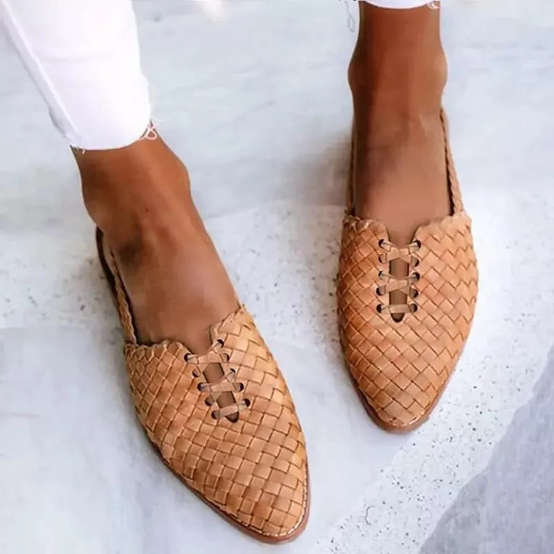 Close-up front view of tan woven leather flats with pointed toe and lace-up detail, worn barefoot on marble flooring