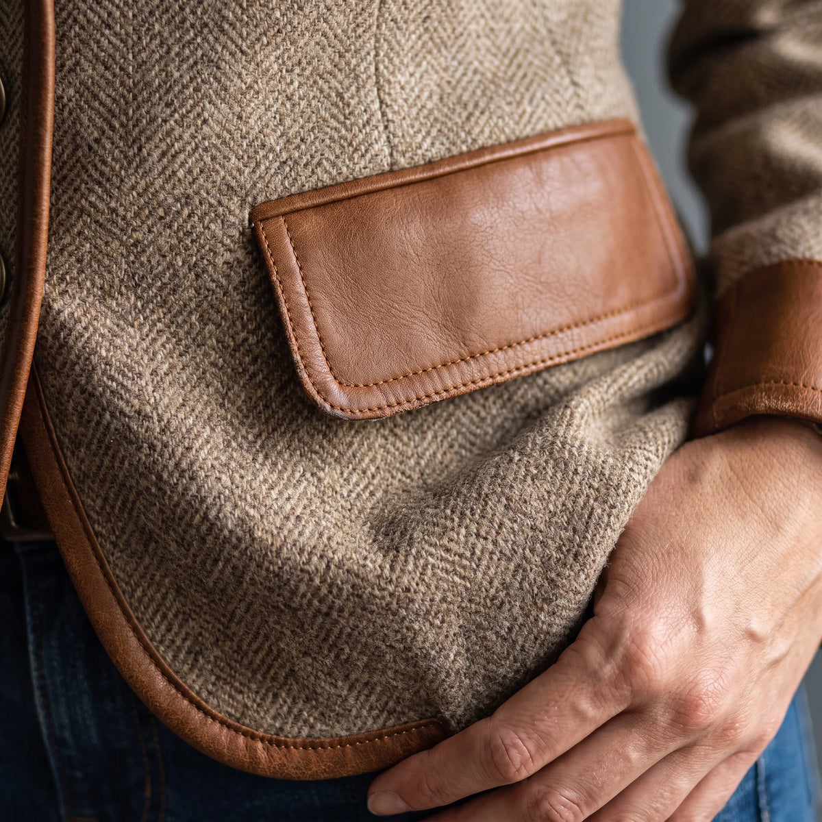 Detail shot of a leather-trimmed flap pocket set against the herringbone tweed body of the jacket