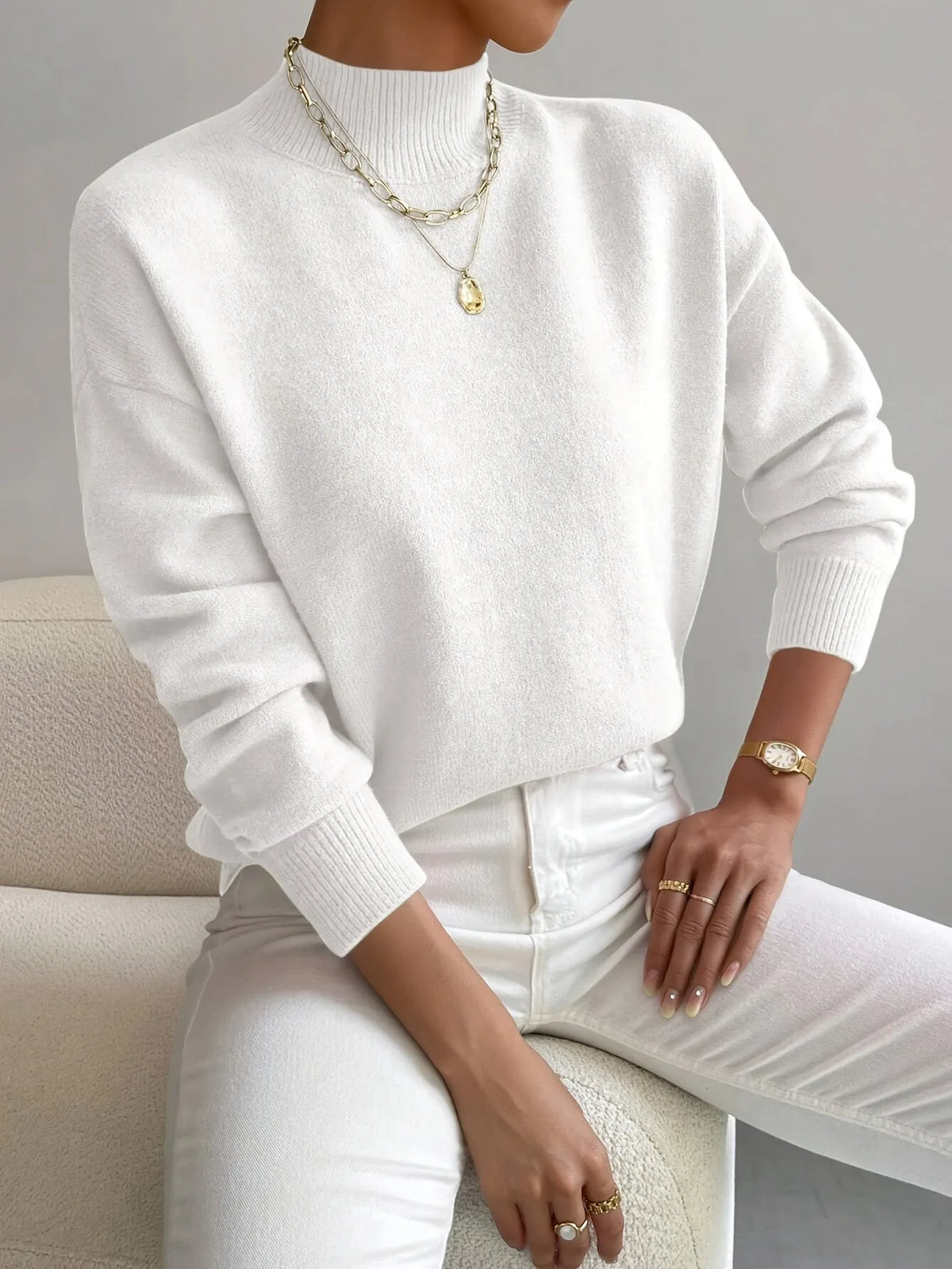 White high neck jumper styled with layered gold necklaces and white trousers, relaxed oversized fit.