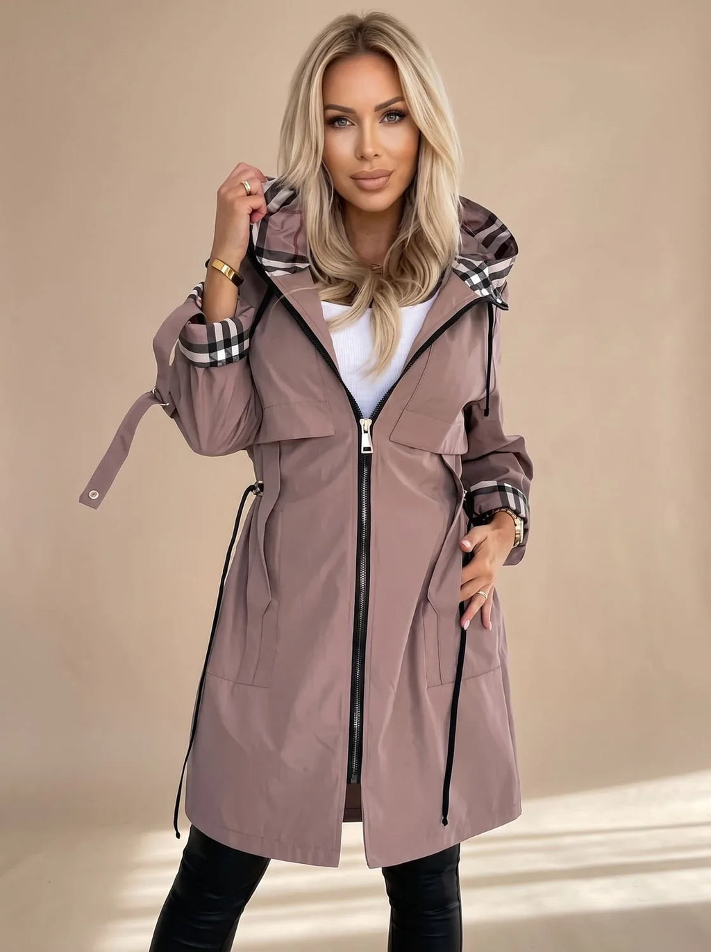 Woman wearing a mocha brown hooded trench coat with checked hood lining, open front and silver zip, styled with black trousers