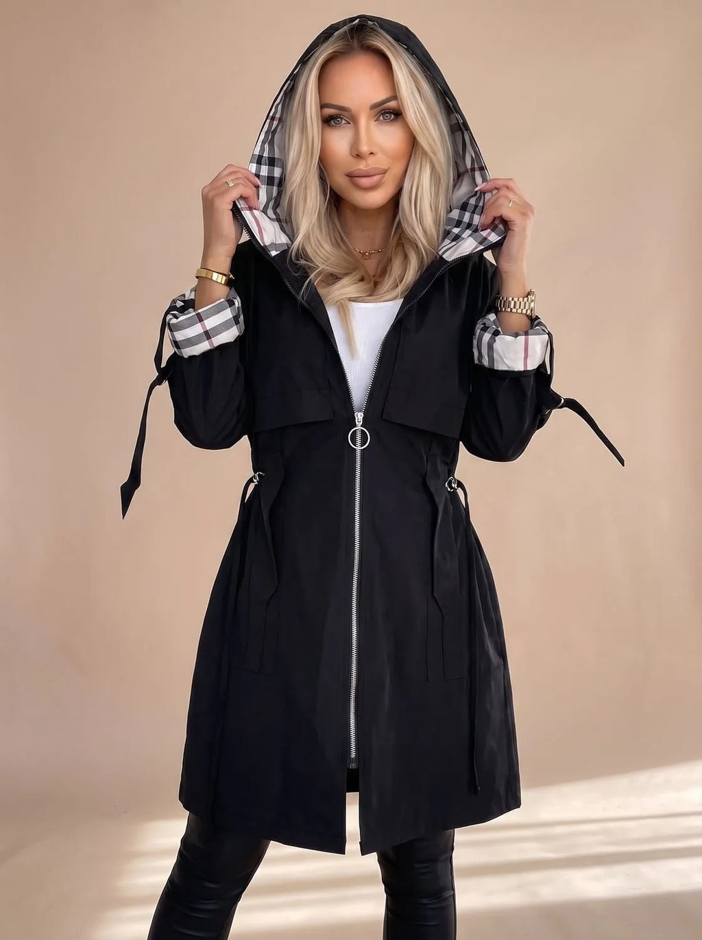 Woman wearing a black hooded trench coat with checked cuff lining, silver ring-pull zip, and drawstring waist detail