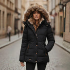 Woman wearing a black hooded puffer jacket with large faux fur trim, zip and button front, on a cobbled street
