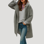Grey hooded longline coat with buttoned front, worn over a grey jumper and jeans, hand raised to hood