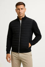 Front view of a man wearing a black quilted jacket with knitted sleeves, stand collar and silver zip, over beige trousers