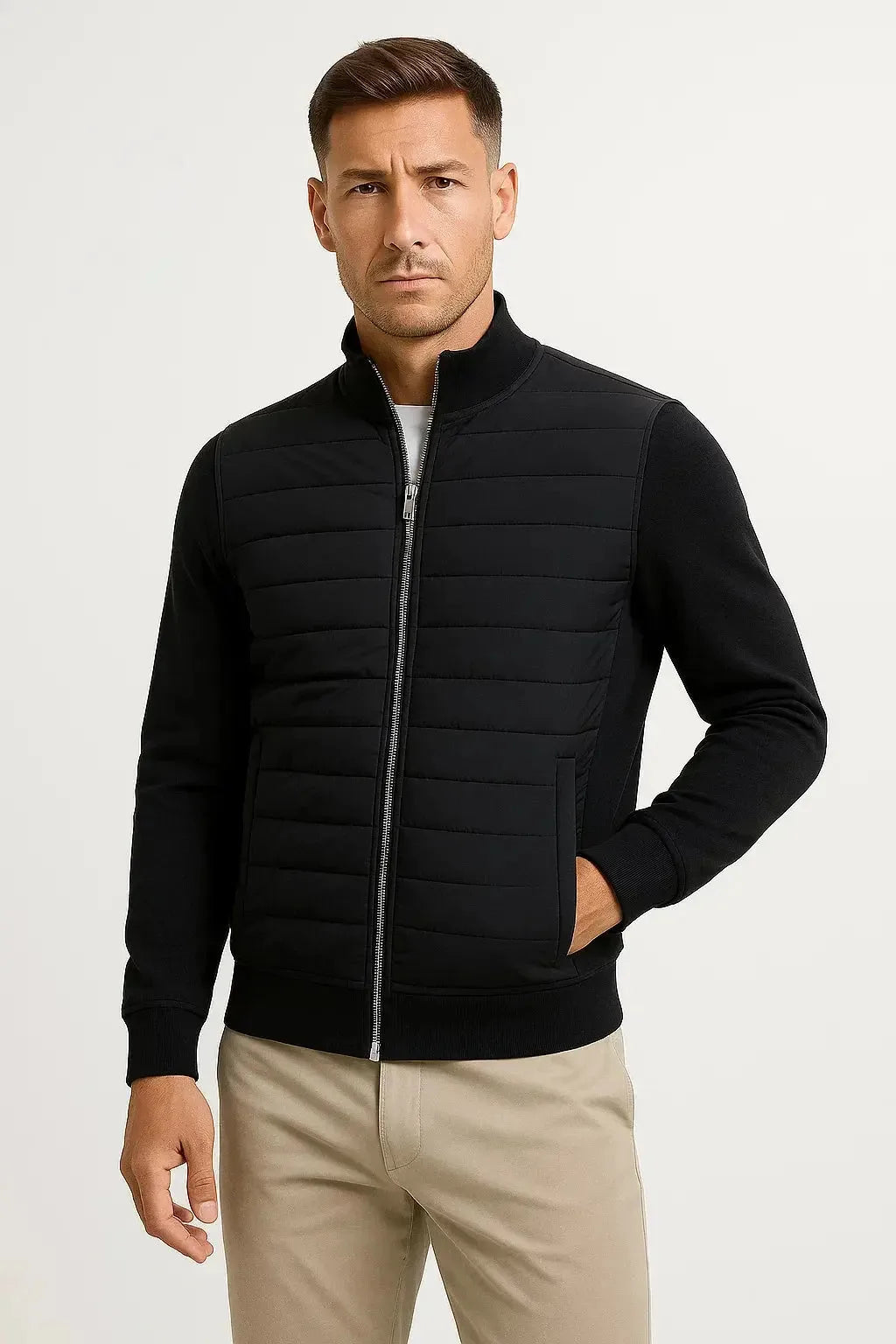Front view of a man wearing a black quilted jacket with knitted sleeves, stand collar and silver zip, over beige trousers