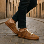 Close-up side profile of a model wearing tan trainers with cream laces, styled with dark jeans on a cobbled street