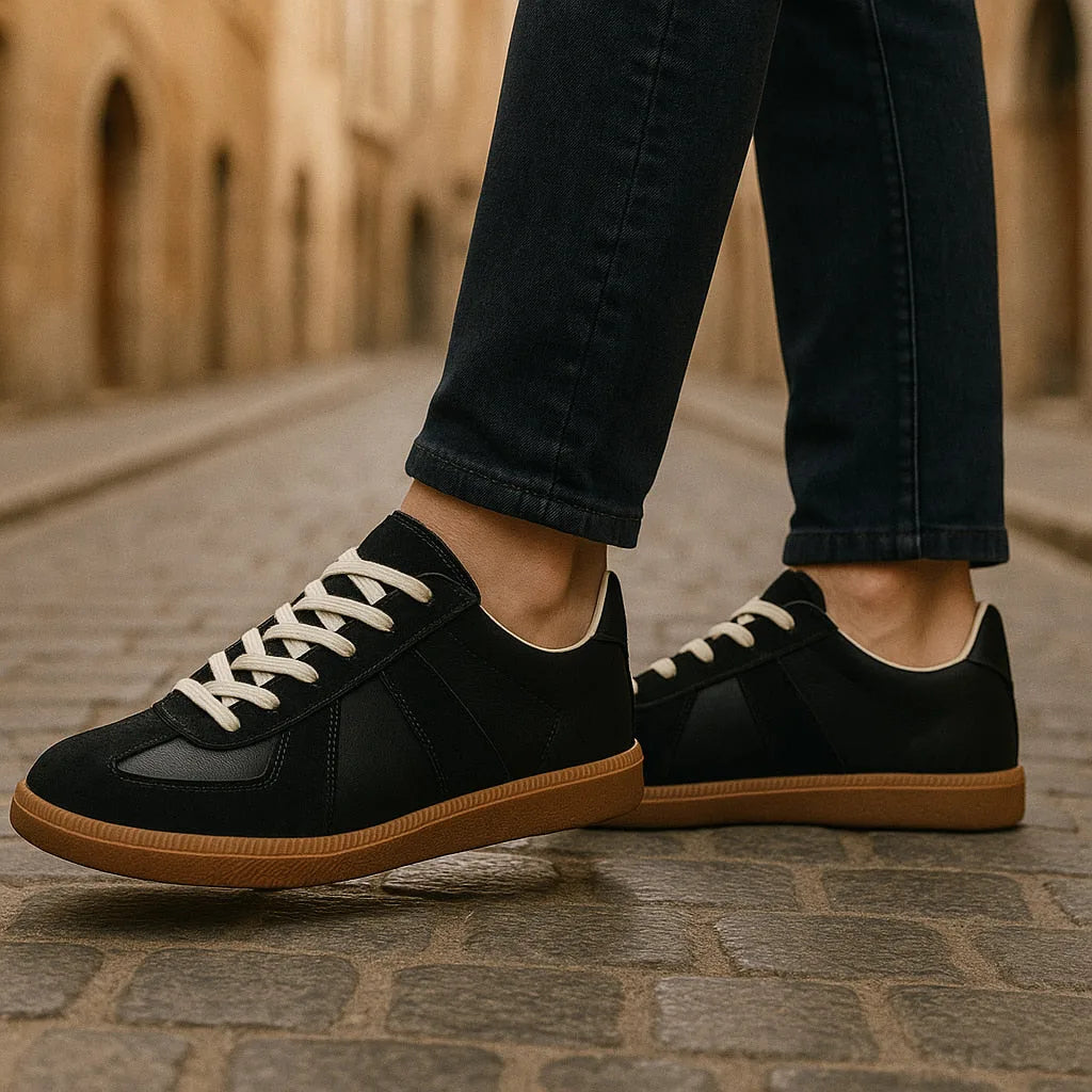 Close-up side profile of black leather and suede trainers worn with dark jeans on a cobbled street