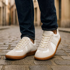 Front-on close-up of off-white suede and leather trainers worn with dark jeans on a cobbled street