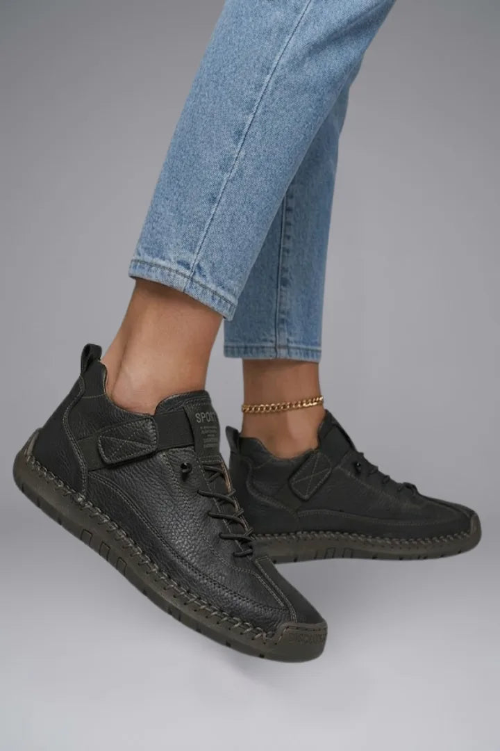 Side view of a model's foot in mid-step wearing black leather trainers with visible EVA sole and stitched trim