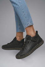 Side view of black leather trainers with elastic strap fastening, shown while worn with rolled denim jeans