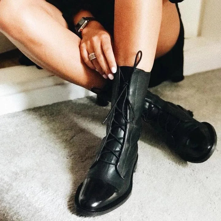 Close-up of a woman lacing up black leather combat boots with a cap toe, seated on stairs