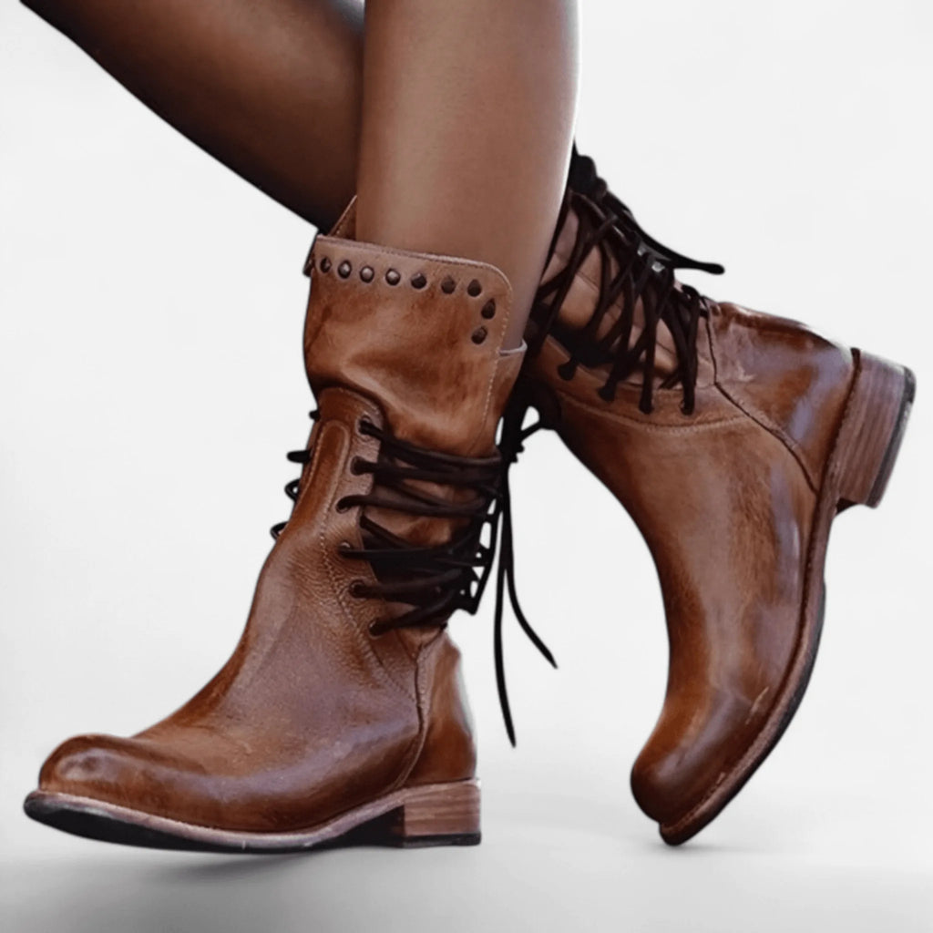 Side view of tan brown leather lace-up boots with stud detailing worn on crossed legs, showing the almond toe shape