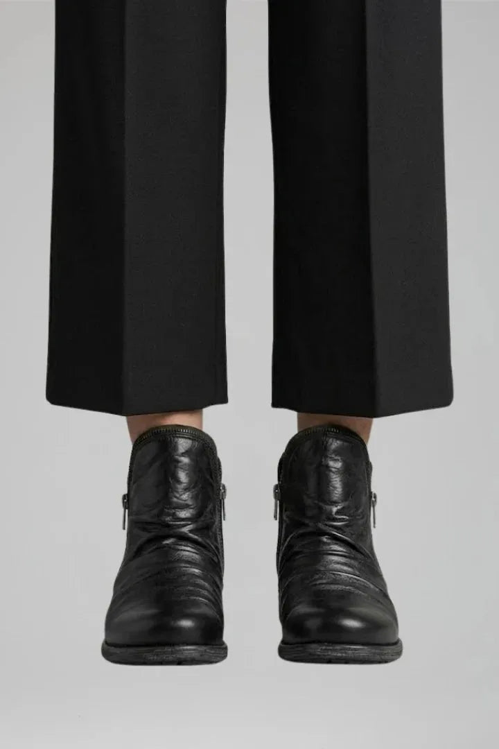 Close-up front view of black ruched leather ankle boots worn with cropped black tailored trousers