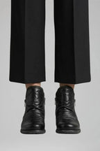 Close-up front view of black ruched leather ankle boots worn with cropped black tailored trousers