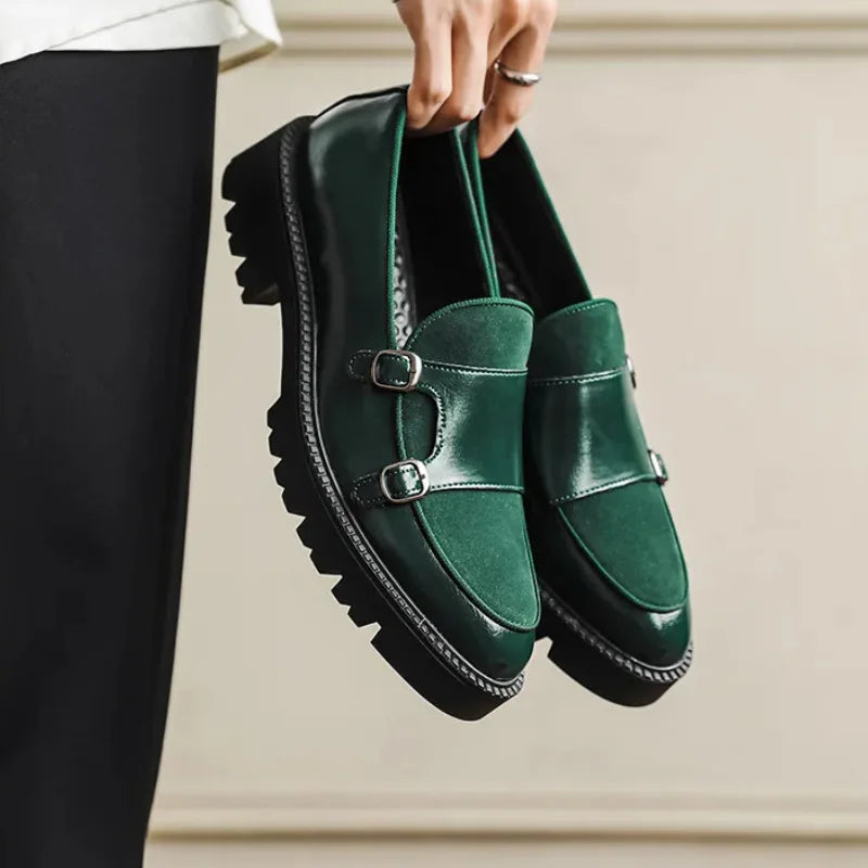 Hand holding a pair of green leather loafers with suede toe panel and twin buckle straps against a neutral wall