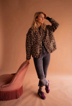 Woman standing beside a pink chair wearing the leopard print bomber jacket fully buttoned with front pockets visible