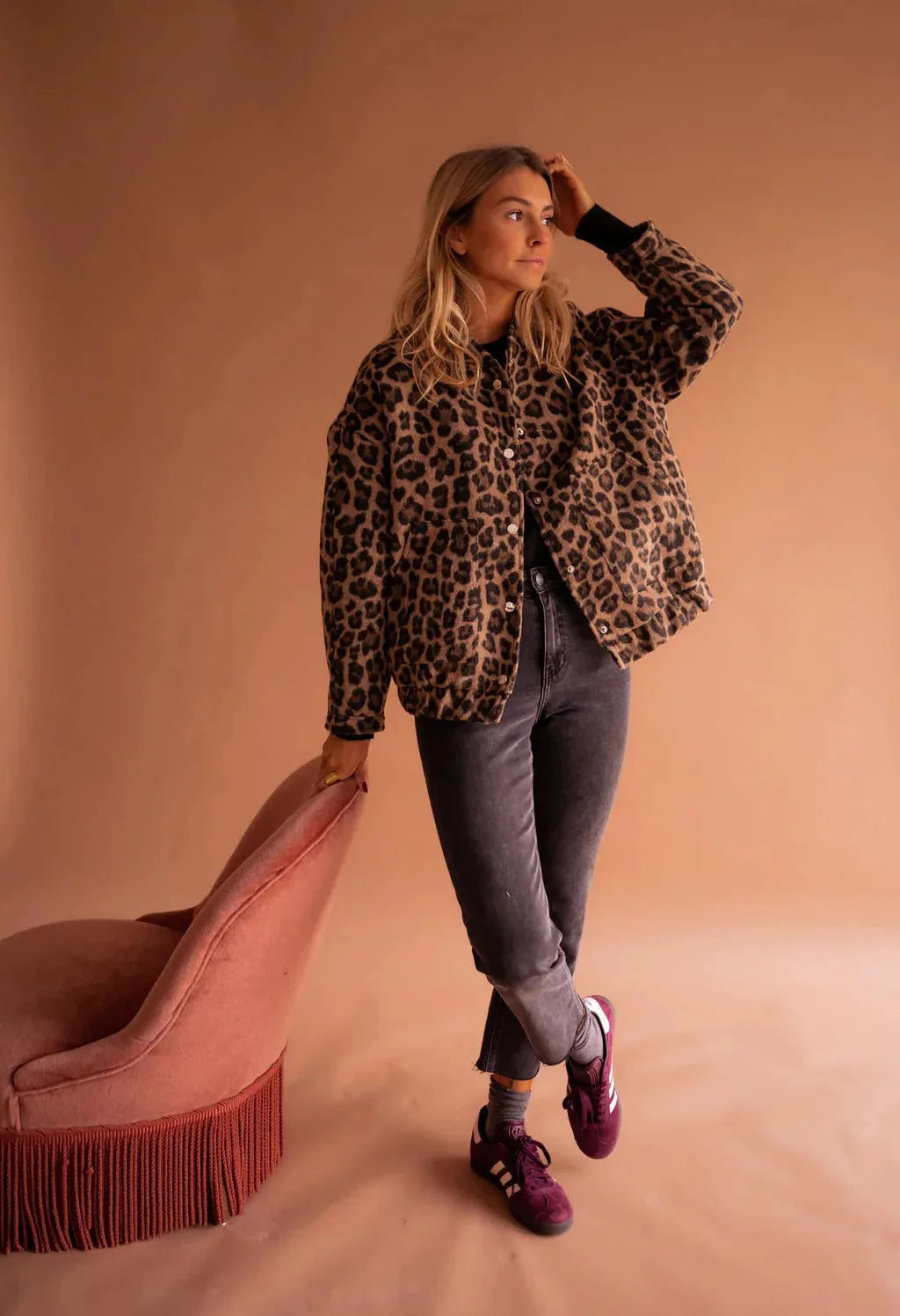 Woman standing beside a pink chair wearing the leopard print bomber jacket fully buttoned with front pockets visible