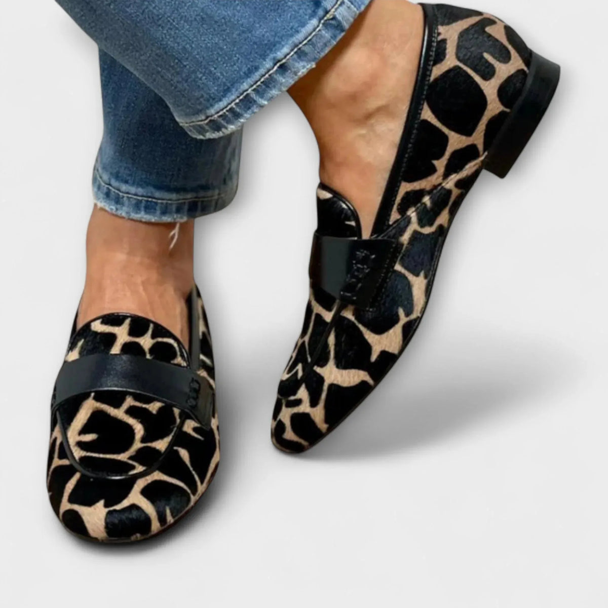 Close-up of a woman's feet wearing leopard-print pony hair loafers with a black leather saddle strap, paired with cropped jeans.