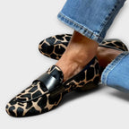 Top-down angled view of leopard-print loafers on crossed feet, showing the pointed toe and black leather trim detail.