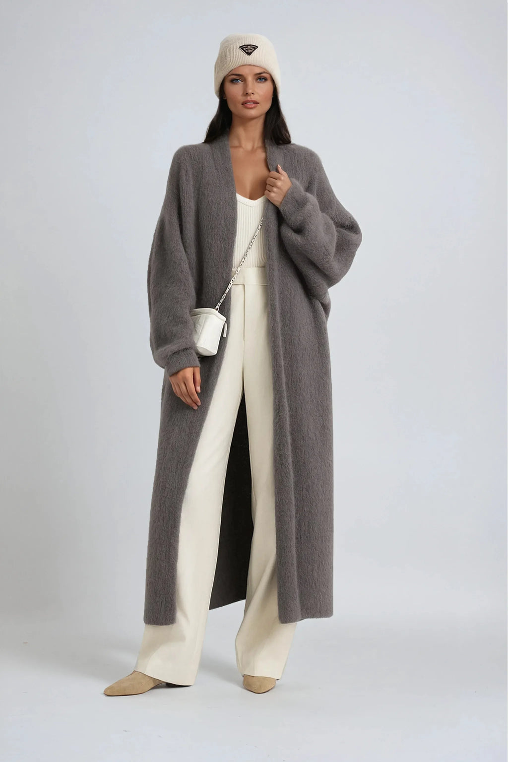 Woman wearing an oversized grey knit long cardigan over cream trousers with a white crossbody bag and knit beanie