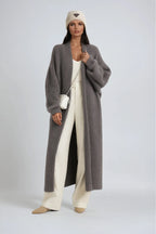 Woman wearing an oversized grey knit long cardigan over cream trousers with a white crossbody bag and knit beanie