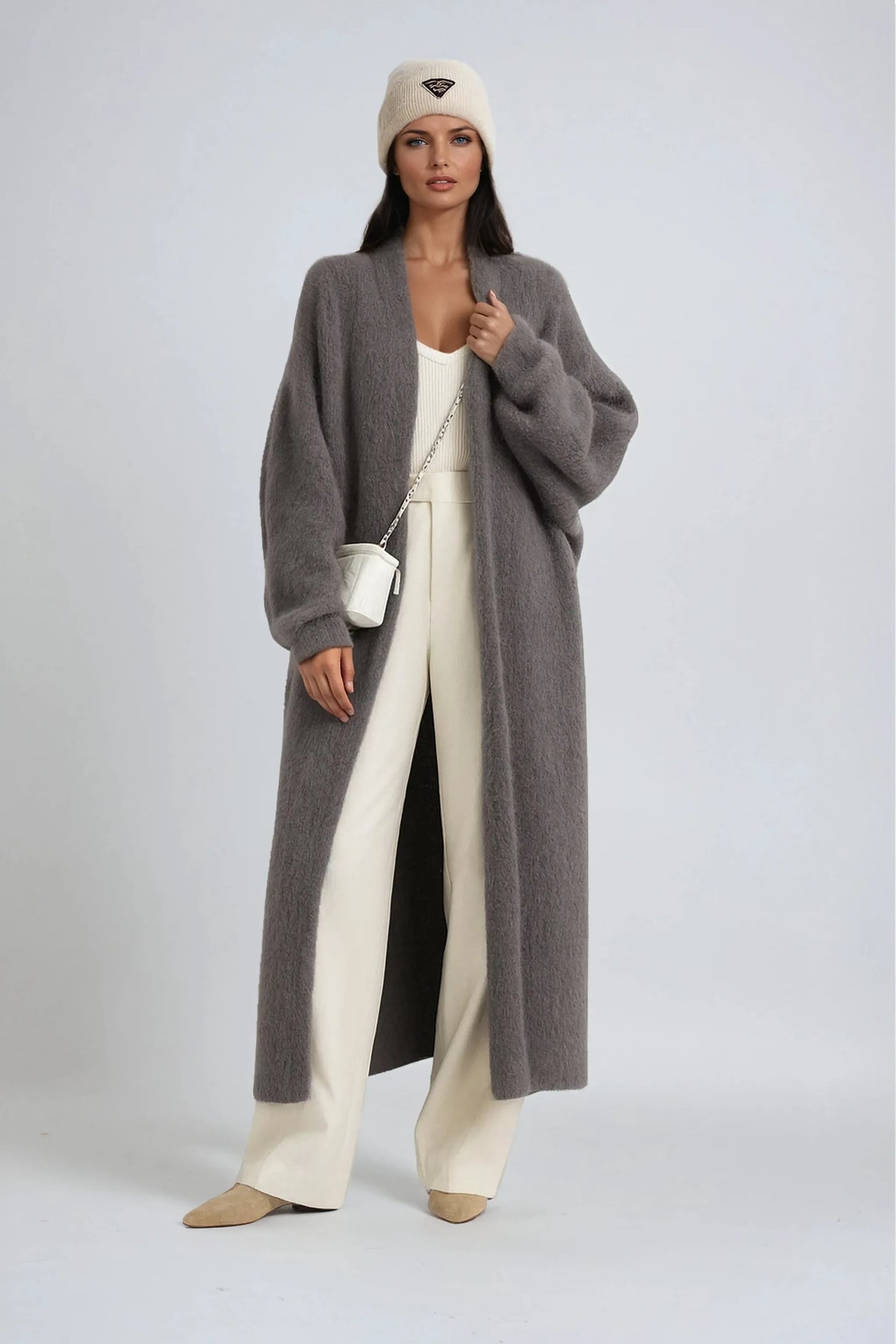 Woman wearing an oversized grey knit long cardigan over cream trousers with a white crossbody bag and knit beanie