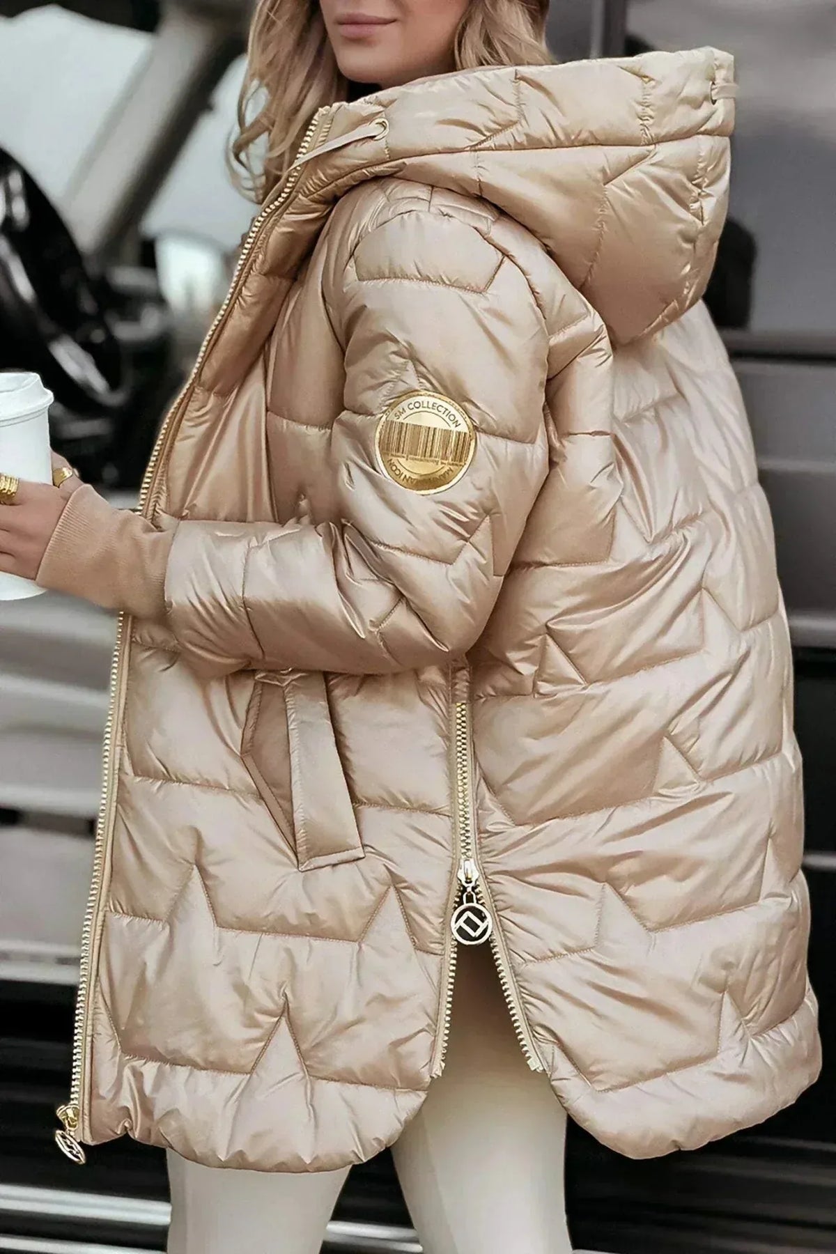 Front view of a champagne gold puffer coat with hood, zip open, holding a white coffee cup
