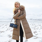 Woman in long brown puffer coat with oversized fur hood holding a black handbag on a snowy landscape